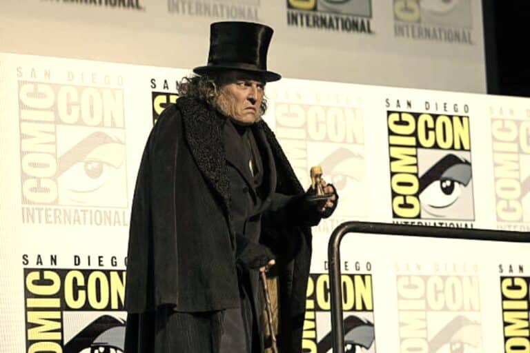 Johnny Depp as Ebenezer Scrooge in costume at Comic-Con, wearing a tall hat and dark cloak, holding a small object, with Comic-Con International branding in the background.