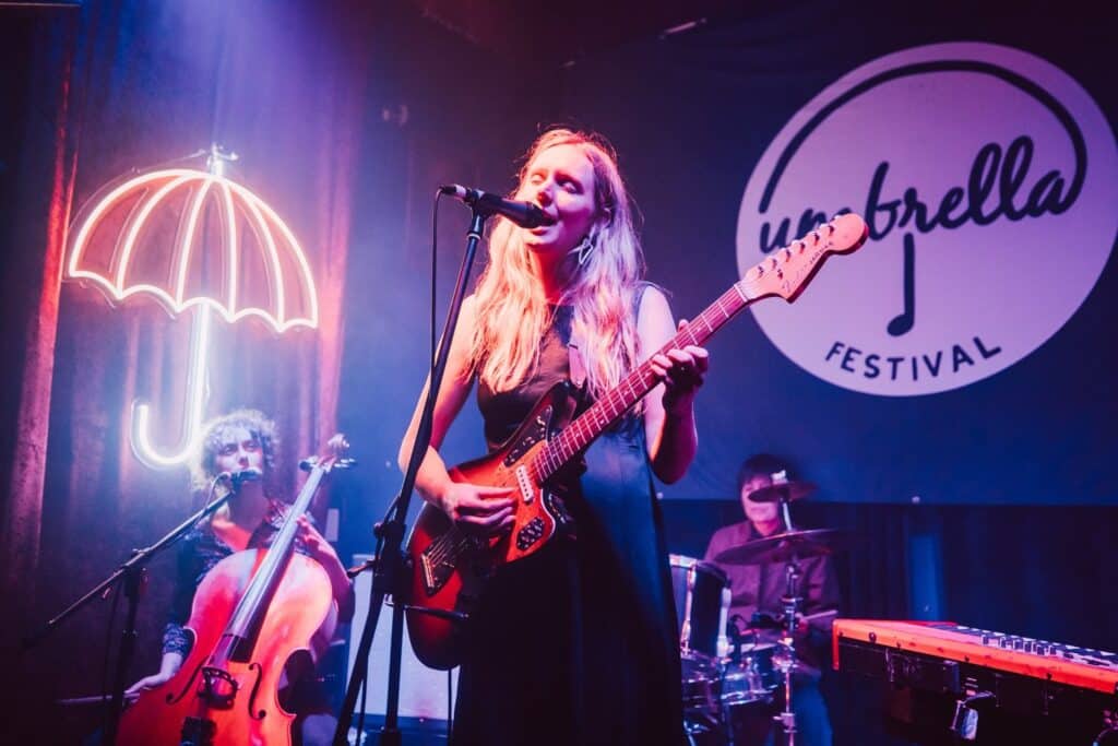 Anya Anastasia singing and playing guitar with cello and drums at Umbrella Festival