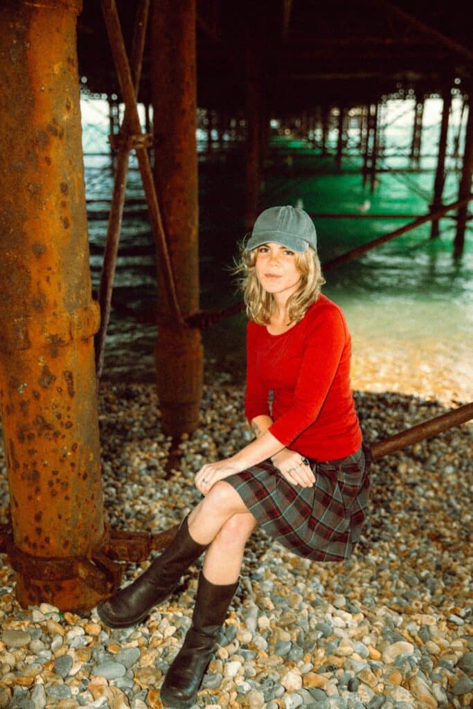 Ella Ion in a red top, checked skirt and grey cap beneath a seaside pier