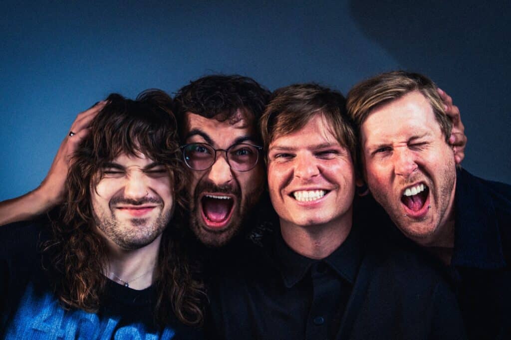The four members of LUMENS close together smiling and pulling faces against a blue backdrop