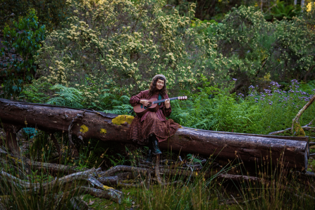 Tasha Zappala playing a ukulele while seated on a fallen tree among ferns