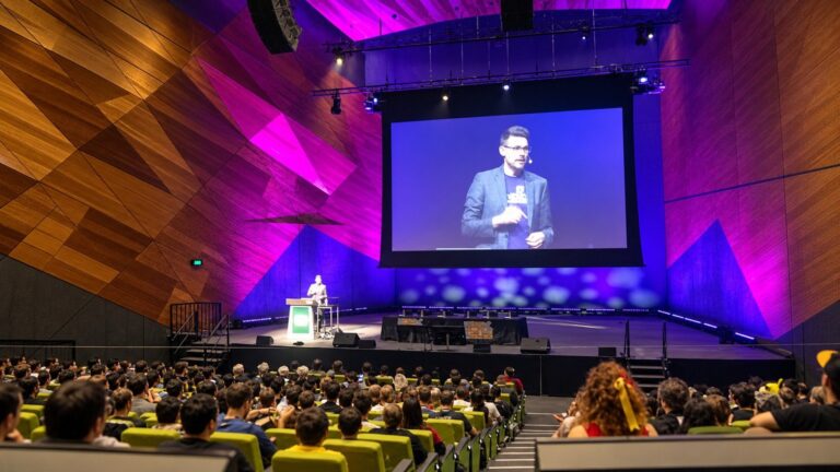 A packed audience watching a presenter on the large screen of a PAX Australia auditorium