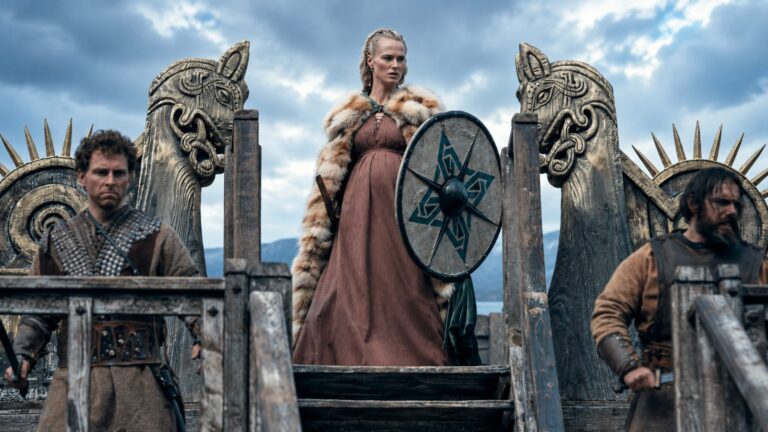 Gunnhild stands with a round shield inside a carved Viking hall in Bloodaxe