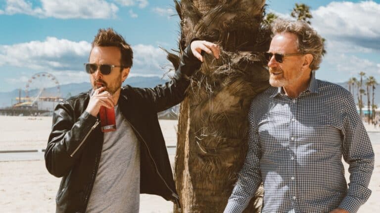 Aaron Paul and Bryan Cranston standing beside a palm tree at the beach