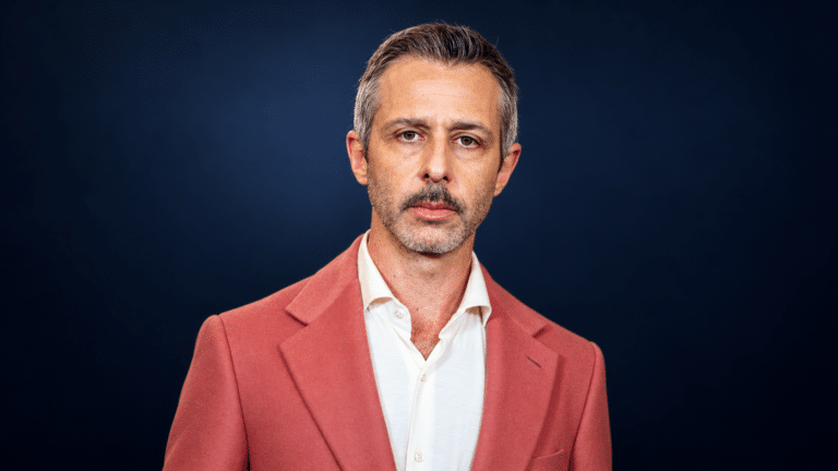 Jeremy Strong in a coral suit against a clean dark background