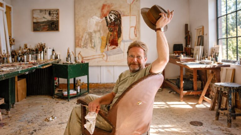 Martin Mull seated in his art studio and raising a hat