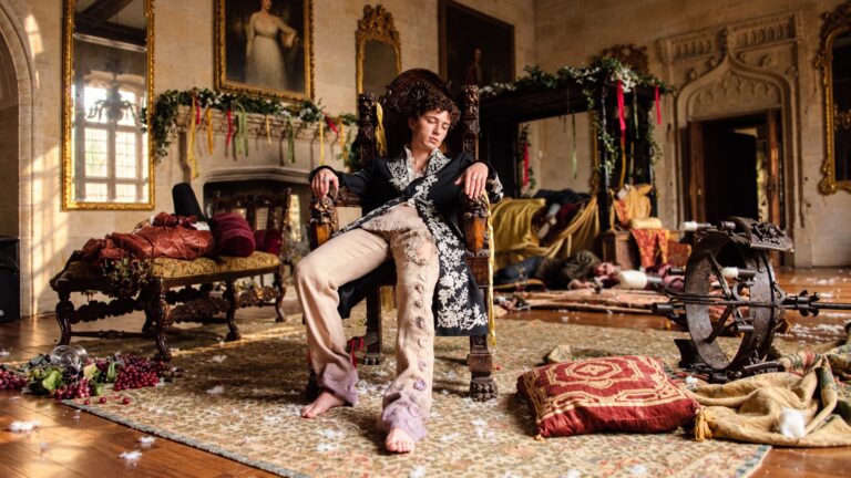 Troye Sivan sits in a period costume inside a disordered castle room